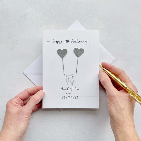 The image shows a beautiful handmade anniversary card with two hearts made from steel coloured card, these are attached by grey yarn, resembling balloons being held by a hand-drawn couple underneath. The text on the card reads "Happy 11th Anniversary" at the top, followed by the couples names and wedding date at the bottom. The card is set on a light background, and the person's hands are seen holding a pen and gently holding the card.
