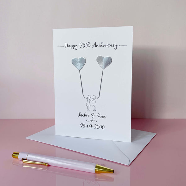 Anniversary card featuring a line drawn couple, each holding a mirror silver heart balloons and personalised text on a pink surface with a pen.