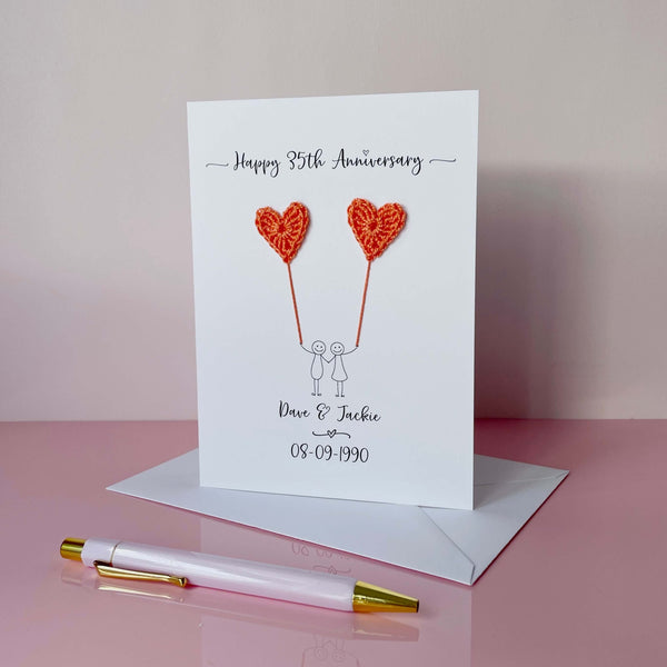 A white card featuring a line drawn couple holding hands and each holding a crochet heart shaped balloon which is made using coral coloured cotton yarn. Happy 35th Anniversary is printed at the top of the card and the couples names and wedding date are printed at the bottom.