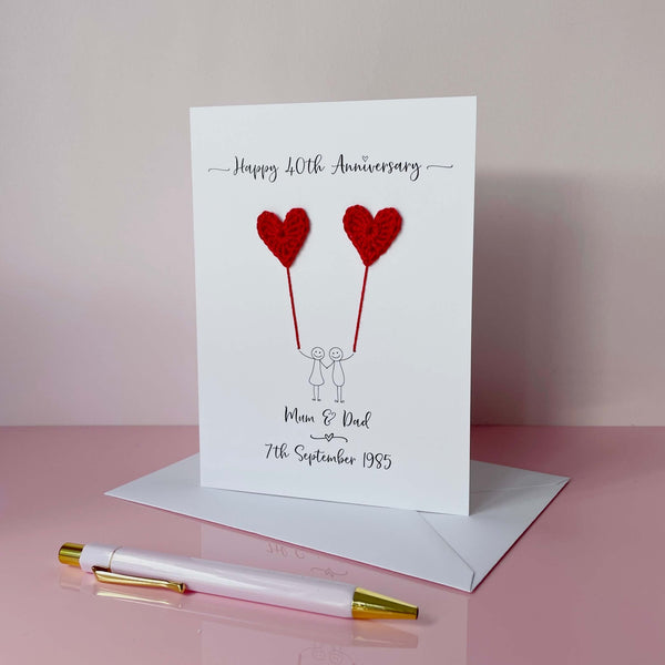 A white card featuring a line drawn couple holding hands and each holding a crochet heart shaped balloon which is made using red coloured cotton yarn. Happy 40th Anniversary is printed at the top of the card and the couples names and wedding date are printed at the bottom.