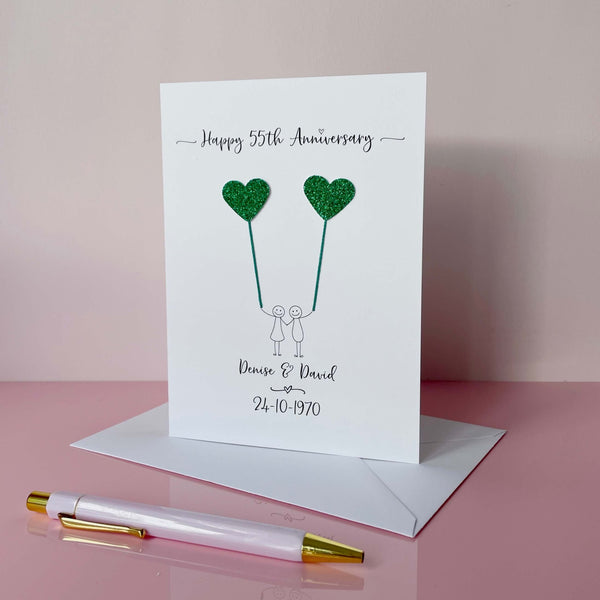 A portrait card with glitter green hearts attached by cotton yarn, resembling balloons being held by a hand-drawn couple underneath. Happy 55th Anniversary is printed at the top, followed by their names and wedding date at the bottom of the card.