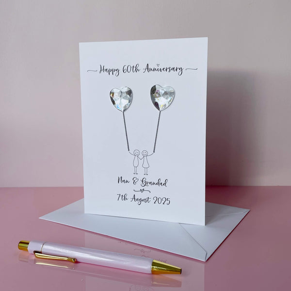 Handmade 60th anniversary card embellished with 2 heart shaped plastic gems, both attached by cotton yarn, resembling balloons being held by a couple underneath. Happy 60th Anniversary is printed at the top, the couples names/ wedding date at the bottom.