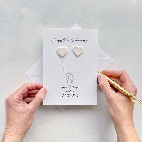 The image shows a beautiful handmade anniversary card with two hearts made from white polymer clay, these are attached by ivory yarn, resembling balloons being held by a hand-drawn couple underneath. The text on the card reads "Happy 9th Anniversary" at the top, followed by the couples names and wedding date at the bottom. The card is set on a light background, and the person's hands are seen holding a pen and gently holding the card.