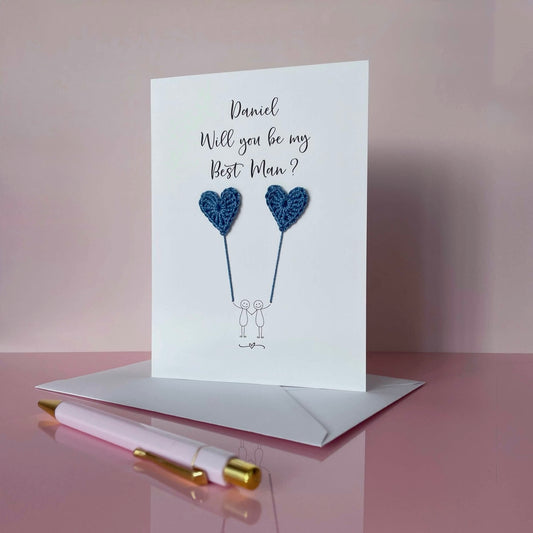 A white card with a line drawn couple printed on it - the couple are each holding a blue crochet heart balloon. Will you be my Best Man is printed at the top of the card with the option of adding a name, making it the perfect keepsake.