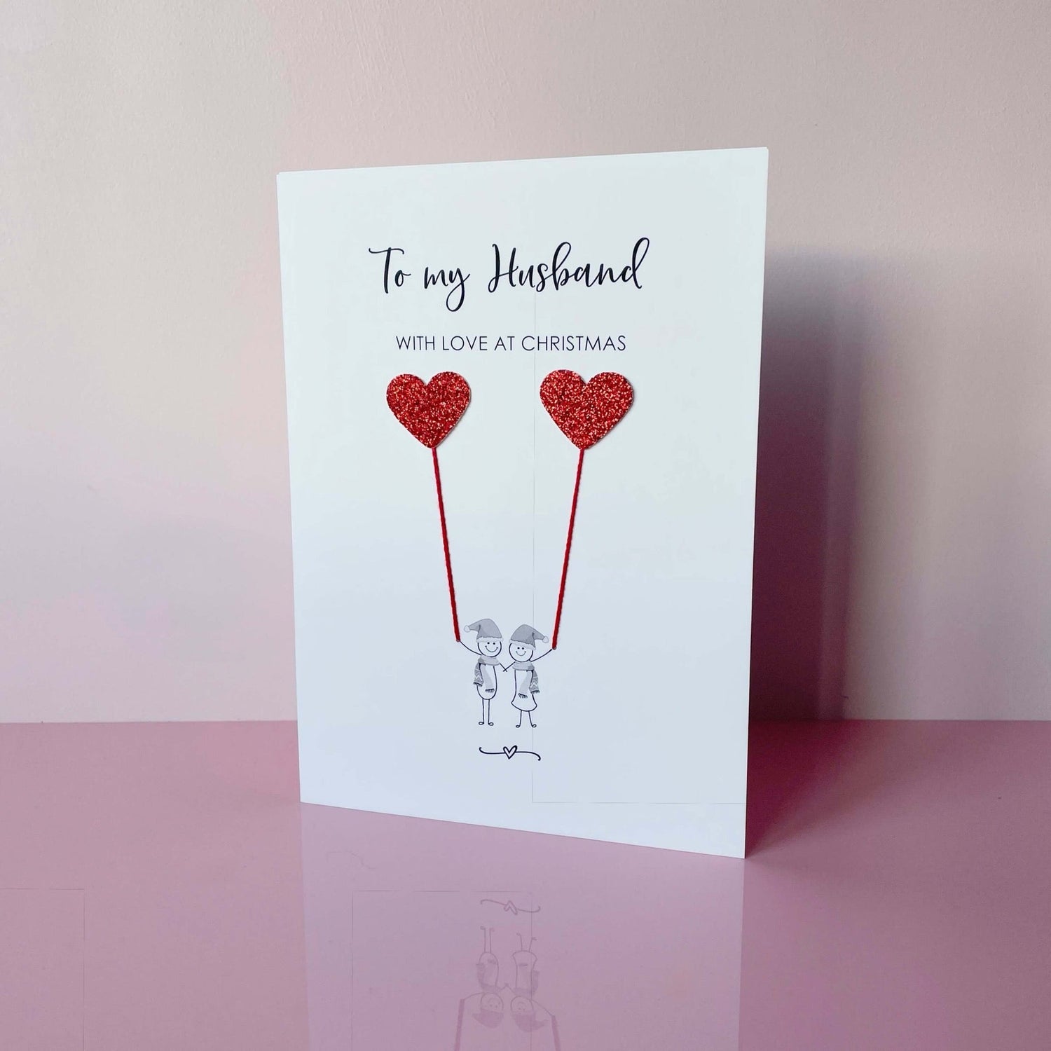 A Christmas card featuring a minimalist design with two figures wearing Santa hats holding red sparkly card heart shaped balloons. The text at the top reads: ‘To my Husband, with love at Christmas’. 