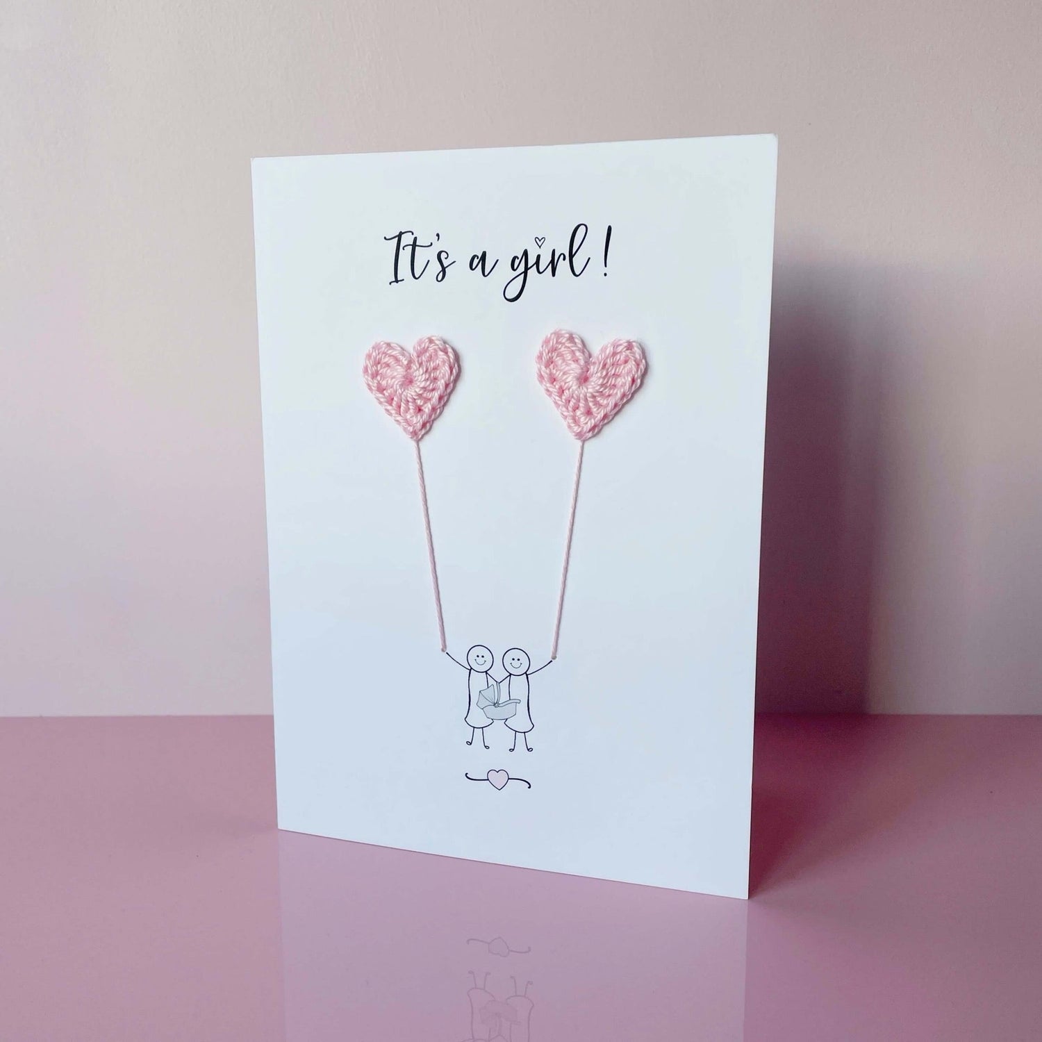A white card featuring a minimalist design with two women figures holding pale pink crochet heart balloons. The text at the top reads: ‘It’s a girl!’. The women are holding a baby seat. The card is photographed on a pale pink background.