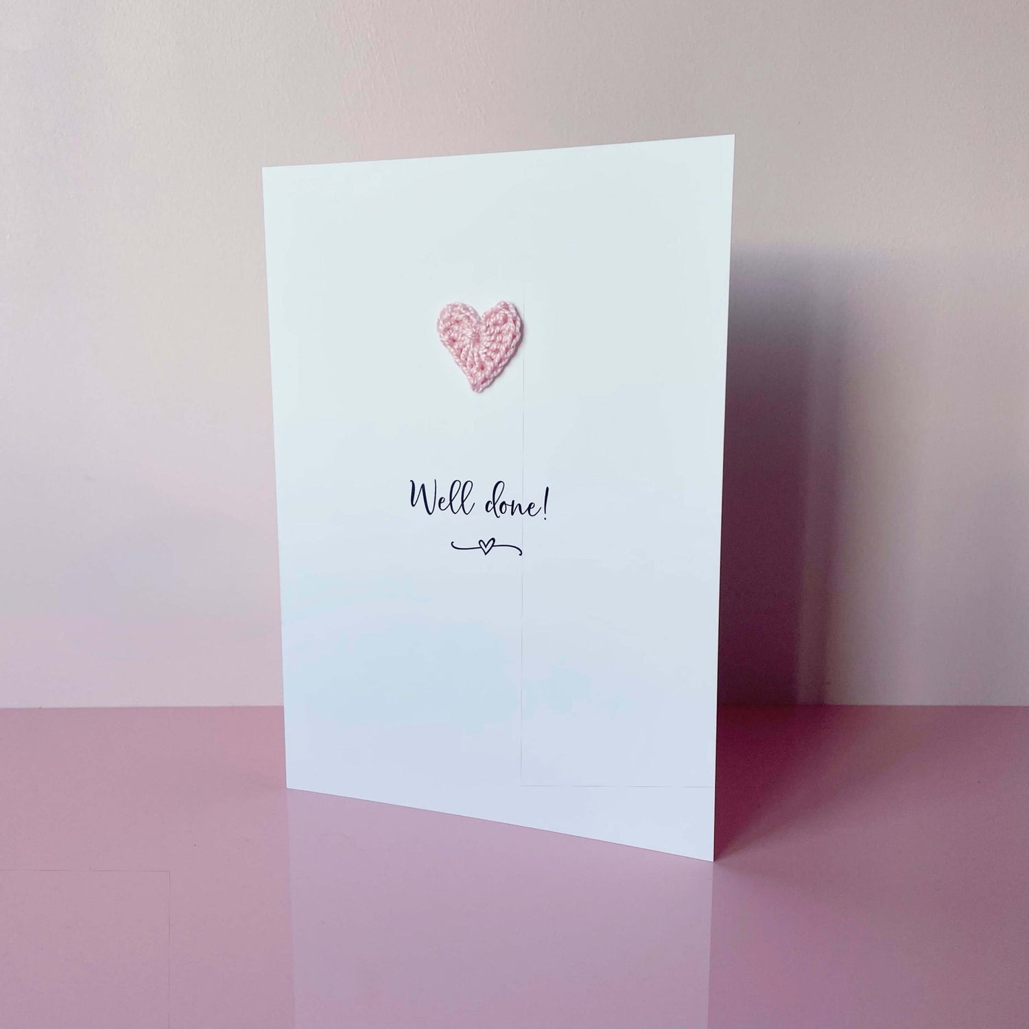 A white portrait card featuring the words ’Well done!’ in elegant black text at the centre. Below the text, a small squiggle and heart design adds a playful touch. Above the words, a pale pink crochet heart is attached, providing a pop of colour