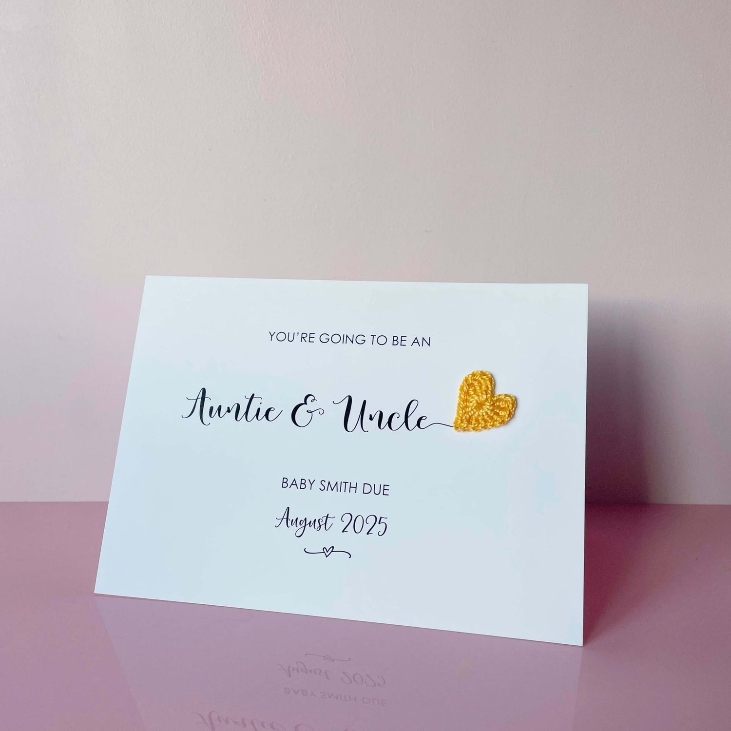 A landscape pregnancy announcement card with the text “You’re going to be an Auntie & Uncle” in elegant black font. Below, it says “Baby Smith due August 2025,” with a small decorative flourish. The card features a small crocheted yellow heart.