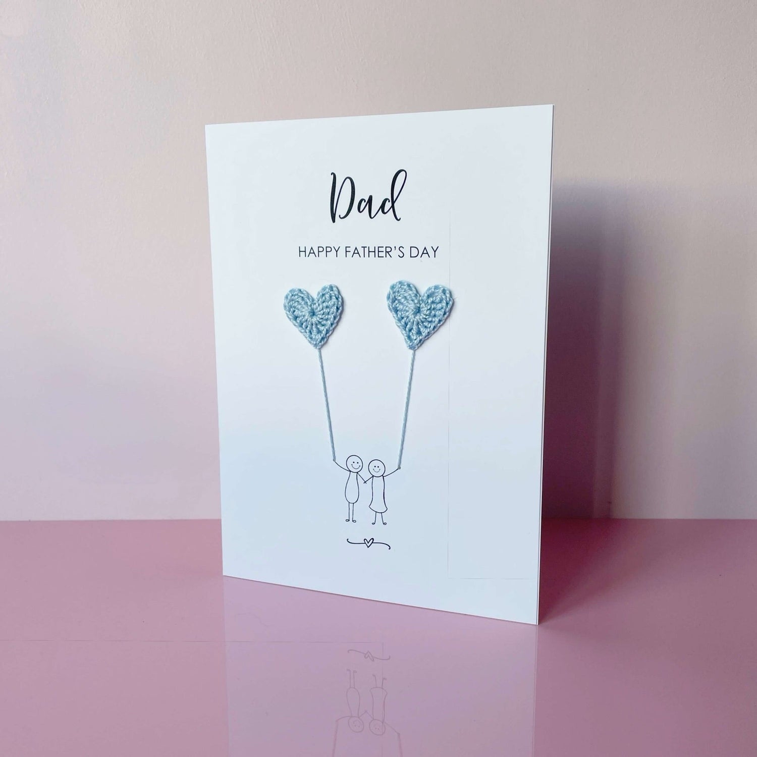 A white Father’s Day card featuring a minimalist design with two line drawn figures holding pale blue crochet heart balloons. The text at the top reads: ‘Dad, Happy Father’s Day. 
