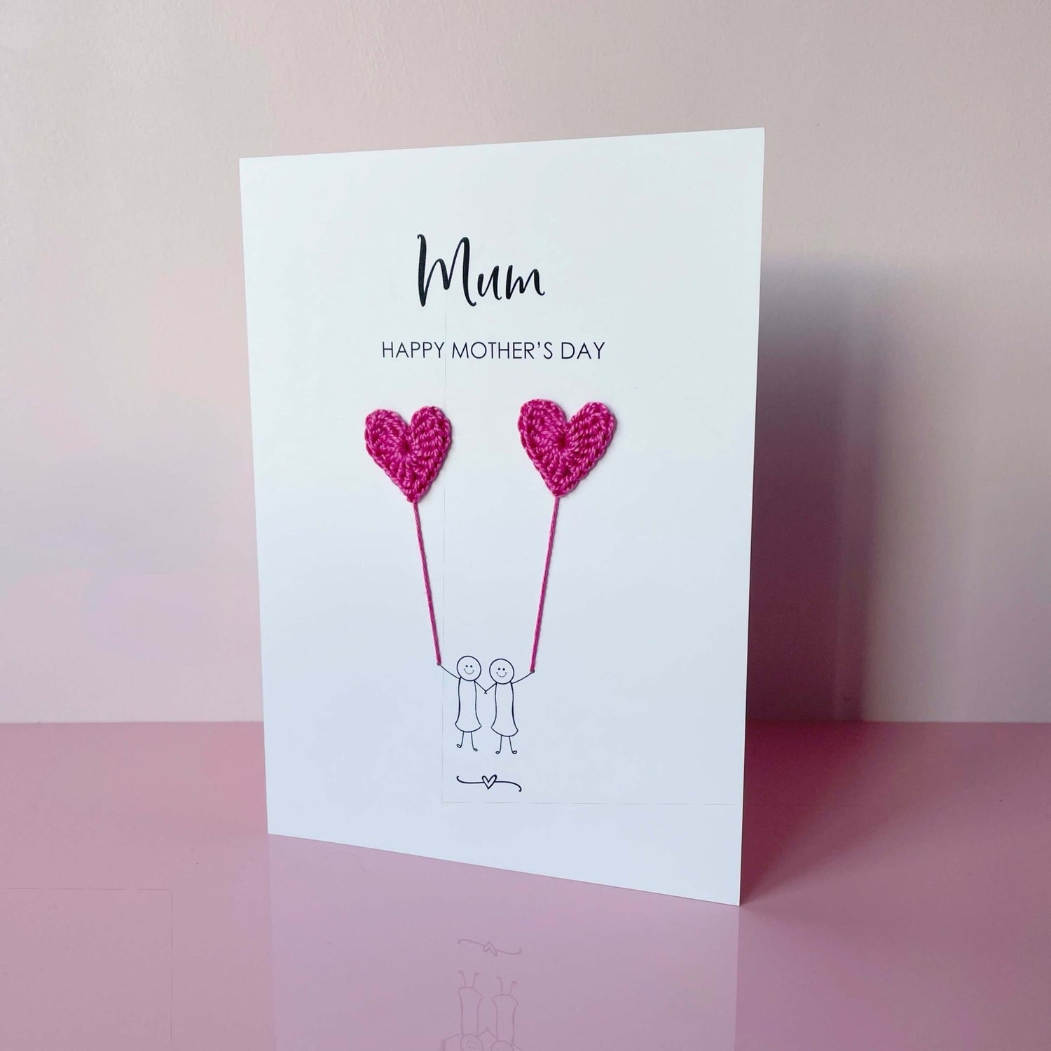 A white Mother’s Day card featuring a minimalist design with two line drawn female figures holding bright pink crochet heart balloons. The text at the top reads: ‘Mum, Happy Mother’s Day. 