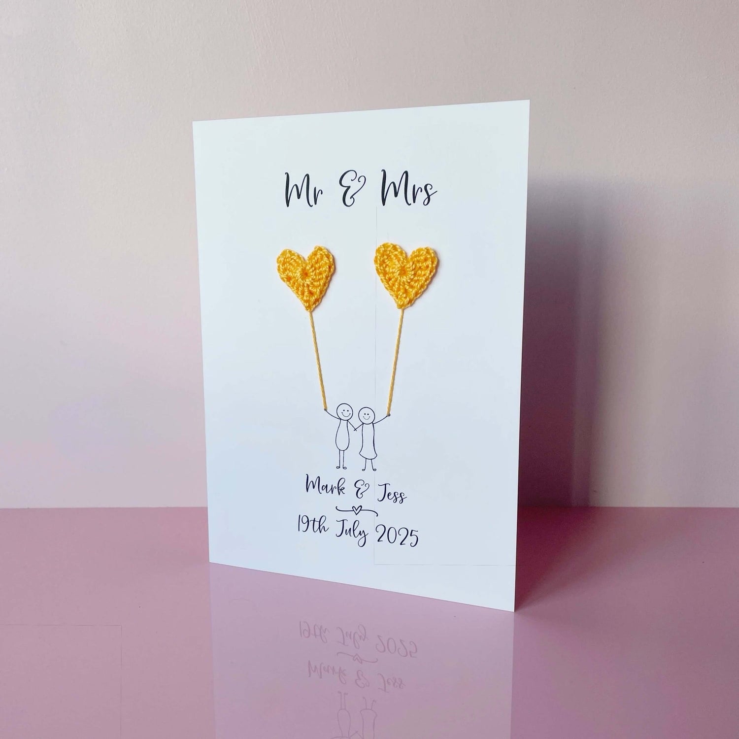 Mr & Mrs is printed at the top of this card in black cursive text.  A line drawn couple holding hands is printed on the card, they are each holding a yellow crochet heart balloon.  Names and wedding date are printed below the couple. 