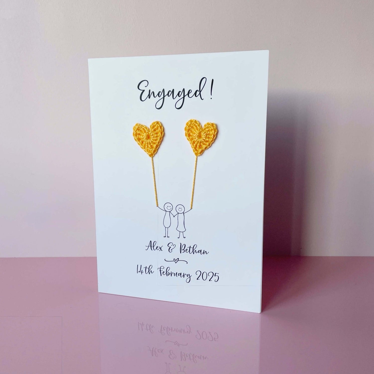 A beautiful handmade engagement card with yellow crochet hearts, these are attached by cotton yarn, resembling balloons being held by a hand-drawn couple underneath. The card reads “Engaged! at the top, followed by the couples names and engagement below. 