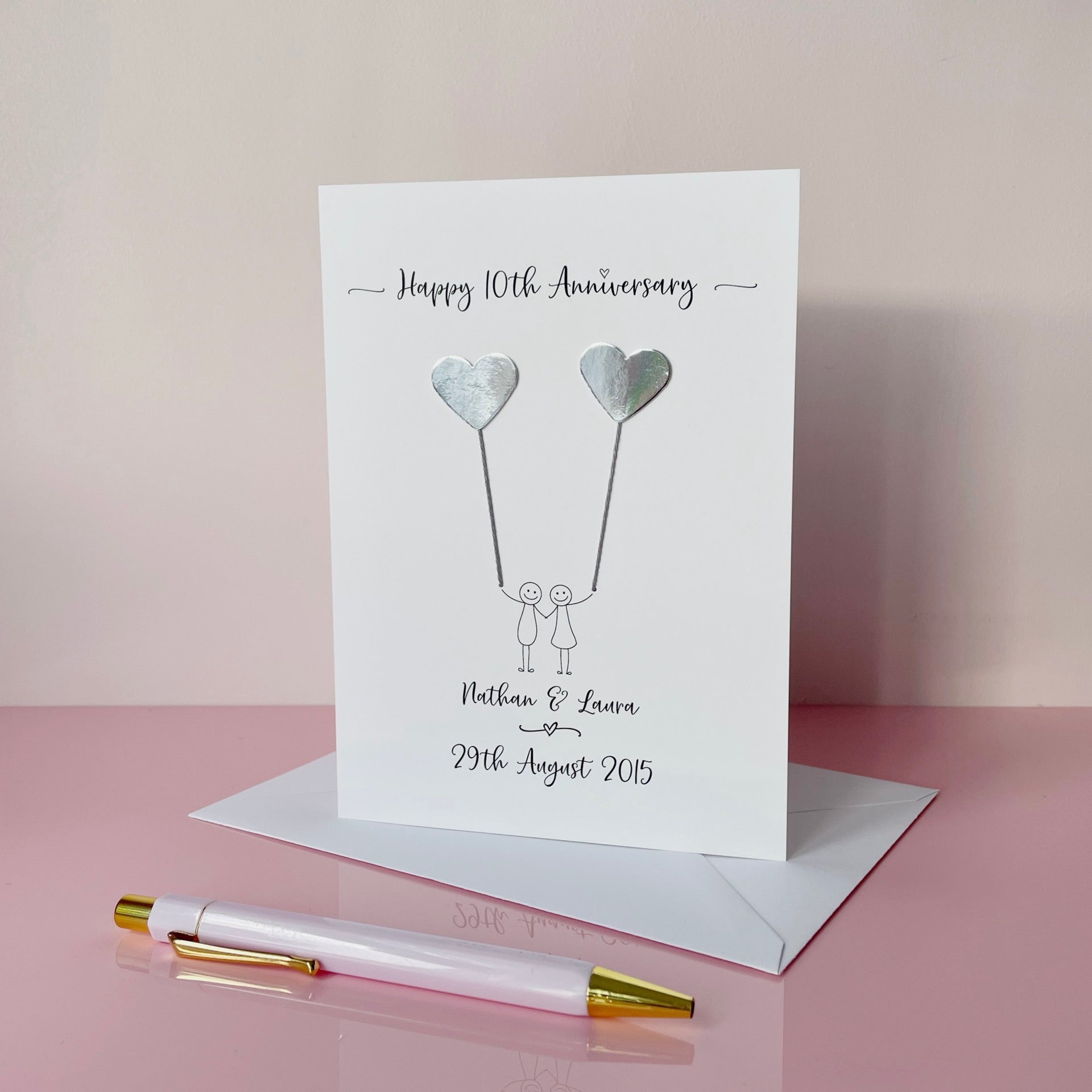 The image shows a beautiful handmade anniversary card with two silver mirror card hearts attached by grey yarn, resembling balloons being held by a hand-drawn couple underneath. The text on the card reads "Happy 10th Anniversary" at the top, followed by the couples names and wedding date at the bottom. 