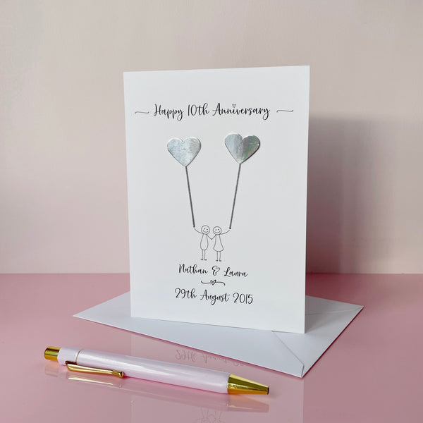 The image shows a beautiful handmade anniversary card with two silver mirror card hearts attached by grey yarn, resembling balloons being held by a hand-drawn couple underneath. The text on the card reads "Happy 10th Anniversary" at the top, followed by the couples names and wedding date at the bottom. 