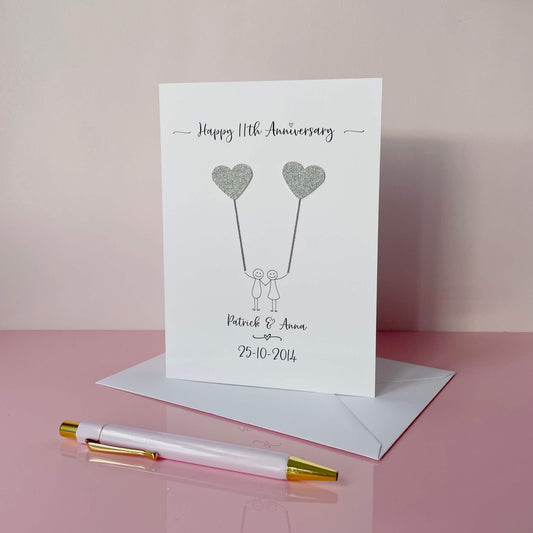 The image shows a beautiful handmade anniversary card with two hearts made from steel coloured card, these are attached by grey yarn, resembling balloons being held by a hand-drawn couple underneath. The text on the card reads "Happy 11th Anniversary" at the top, followed by the couples names and wedding date at the bottom. The card is set on a pink background.