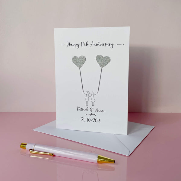 The image shows a beautiful handmade anniversary card with two hearts made from steel coloured card, these are attached by grey yarn, resembling balloons being held by a hand-drawn couple underneath. The text on the card reads "Happy 11th Anniversary" at the top, followed by the couples names and wedding date at the bottom. The card is set on a pink background.