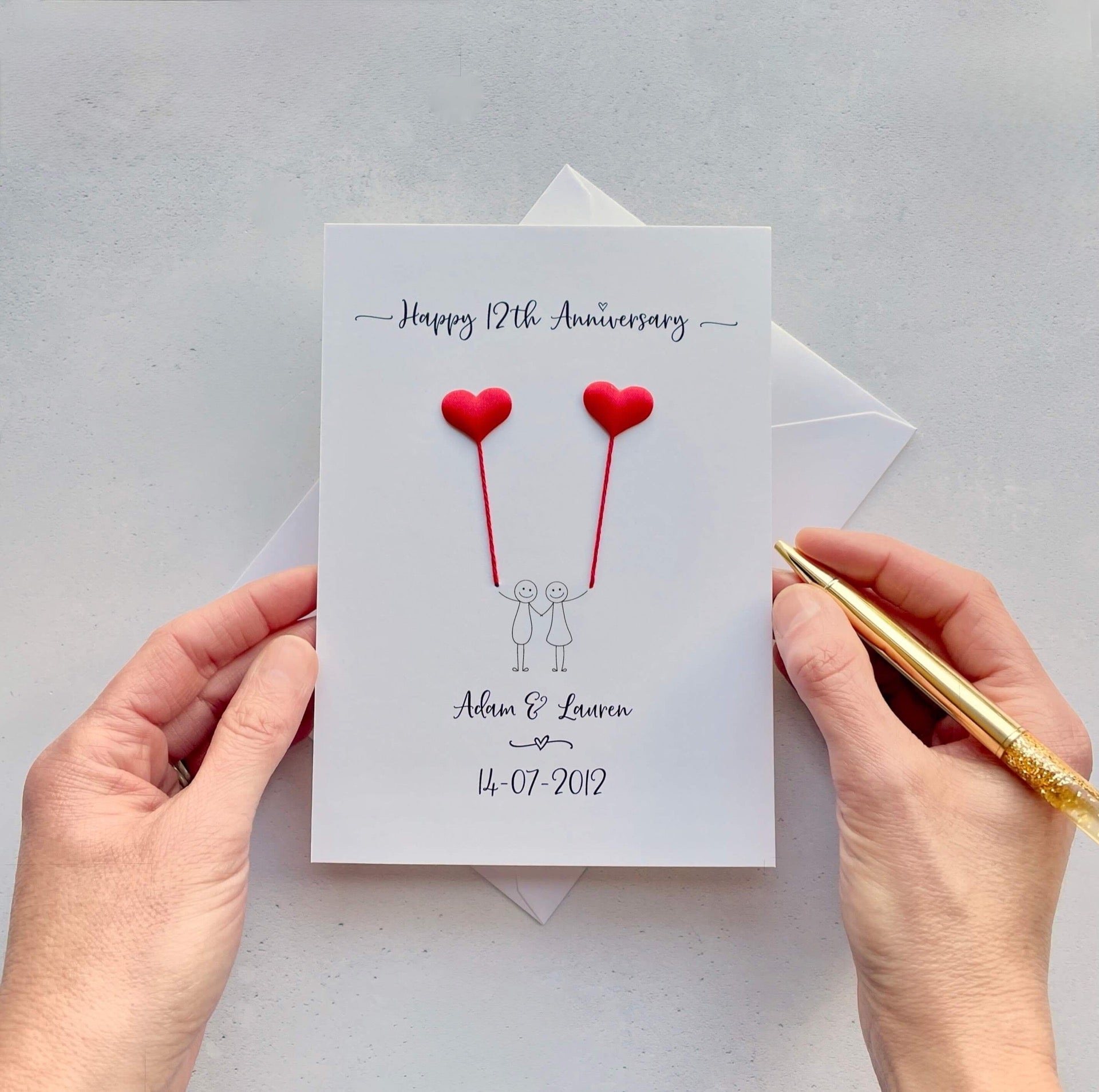 The image shows a beautiful handmade anniversary card with small red silk padded hearts, these are attached by red yarn, resembling balloons being held by a hand-drawn couple underneath. The text on the card reads "Happy 12th Anniversary" at the top, followed by the couples names and wedding date at the bottom. The card is set on a light background, and the person's hands are seen holding a pen and gently holding the card.