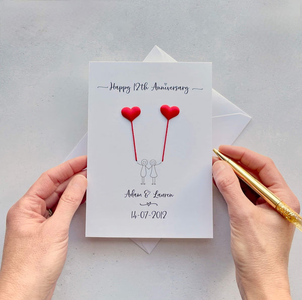 The image shows a beautiful handmade anniversary card with small red silk padded hearts, these are attached by red yarn, resembling balloons being held by a hand-drawn couple underneath. The text on the card reads "Happy 12th Anniversary" at the top, followed by the couples names and wedding date at the bottom. The card is set on a light background, and the person's hands are seen holding a pen and gently holding the card.