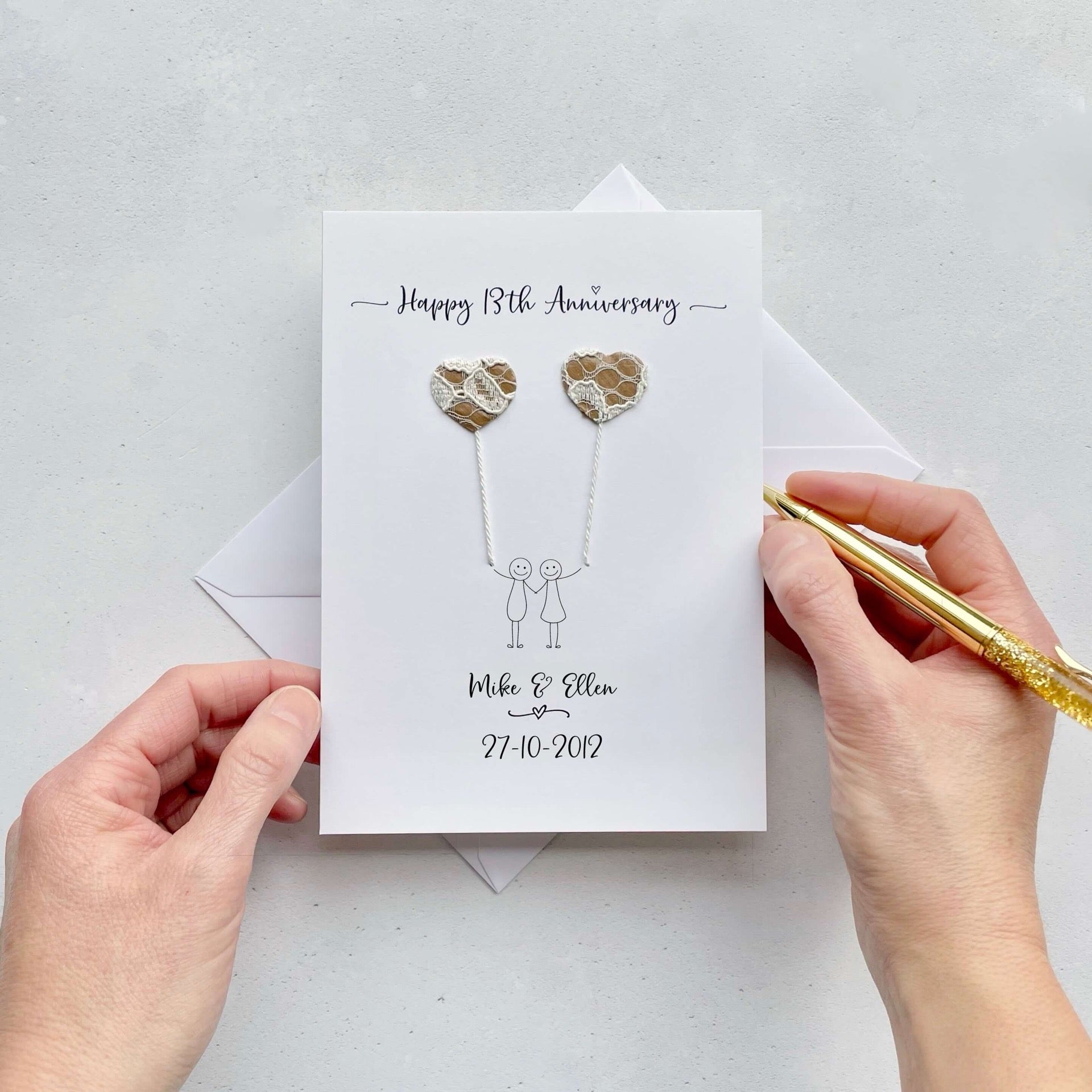 The image shows a beautiful handmade anniversary card with two hearts made from genuine lace and kraft paper, these are attached by ivory yarn, resembling balloons being held by a hand-drawn couple underneath. The text on the card reads "Happy 13th Anniversary" at the top, followed by the couples names and wedding date at the bottom.