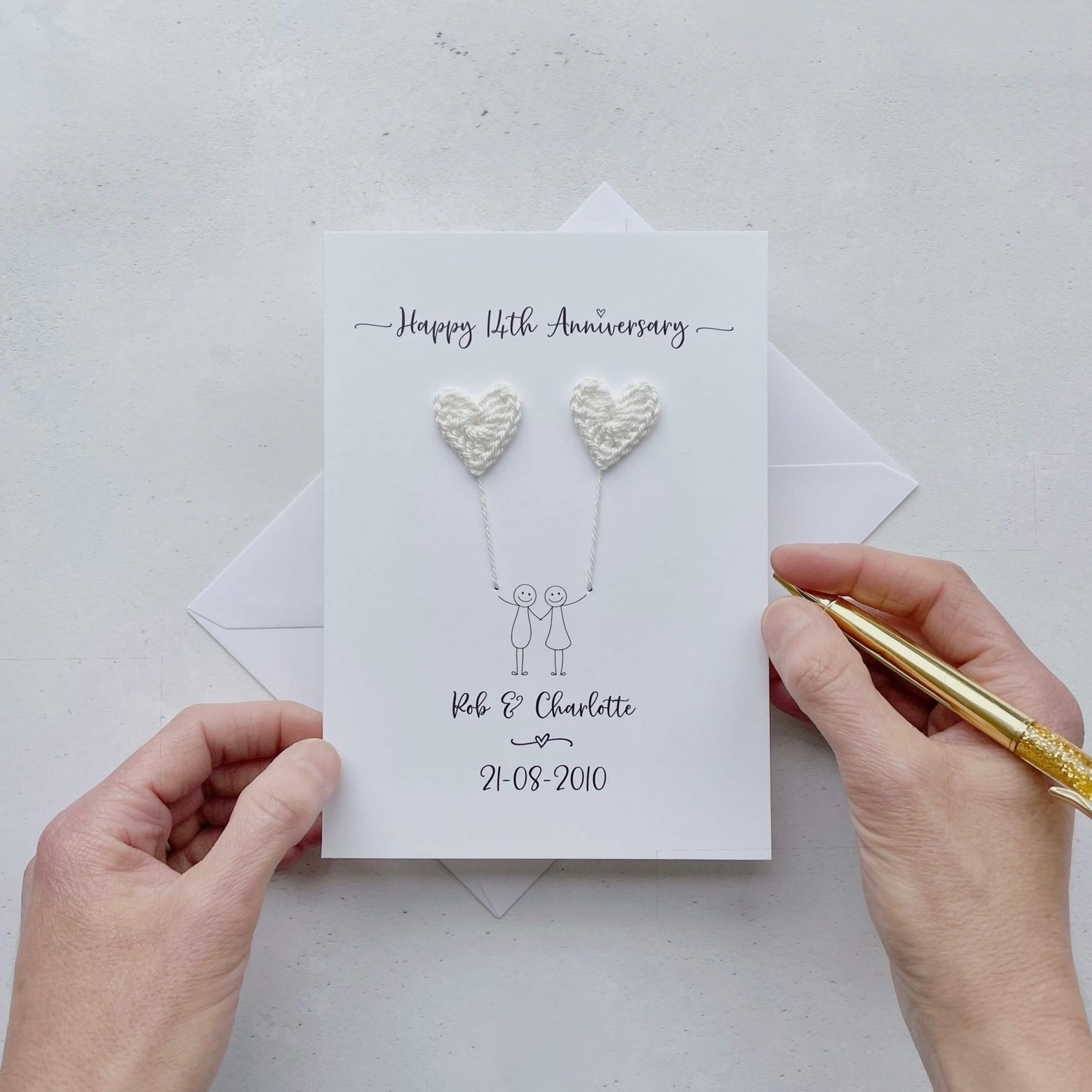 The image shows a beautiful handmade anniversary card with ivory coloured crochet hearts, these are attached by ivory yarn, resembling balloons being held by a hand-drawn couple underneath. The text on the card reads "Happy 14th Anniversary" at the top, followed by the couples names and wedding date at the bottom.