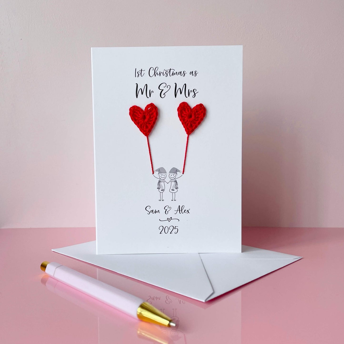 A white card featuring a line drawn couple holding hands and each holding a red crochet heart balloon. ‘1st Christmas as Mr & Mrs’ is printed at the top of the card and the couples names and the year are printed just below the couple. The couple are drawn wearing winter scarves and santa hats.