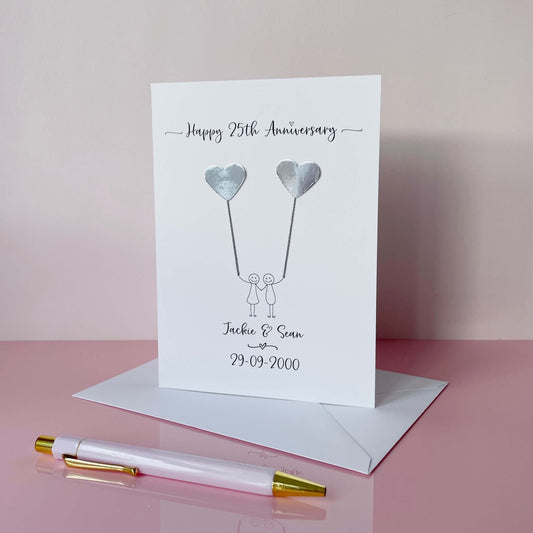 Anniversary card featuring a line drawn couple, each holding a mirror silver heart balloons and personalised text on a pink surface with a pen.