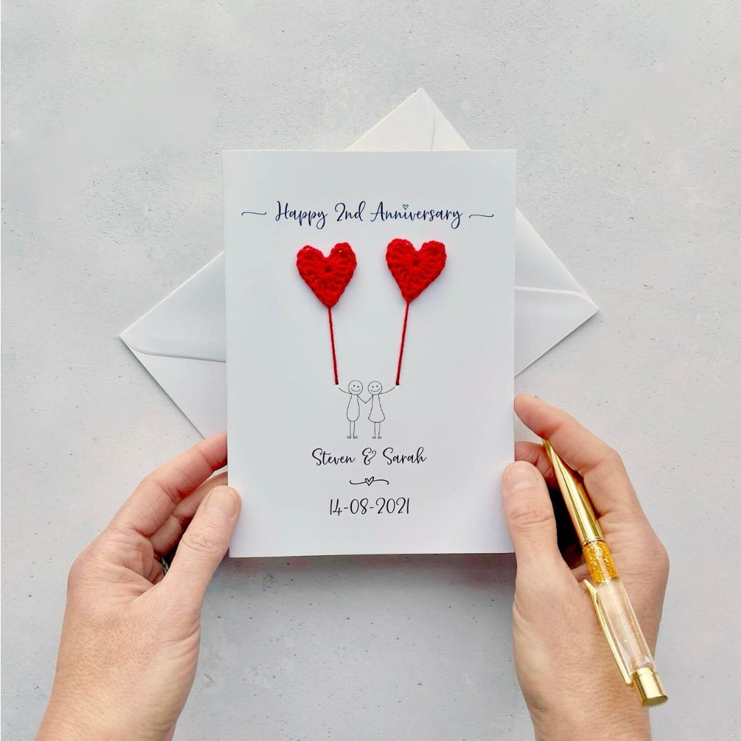 A white card featuring a line drawn couple each holding a red crochet heart made from cotton yarn. Happy 2nd Anniversary is printed at the top of the card and the couples names and wedding date are printed at the bottom. 