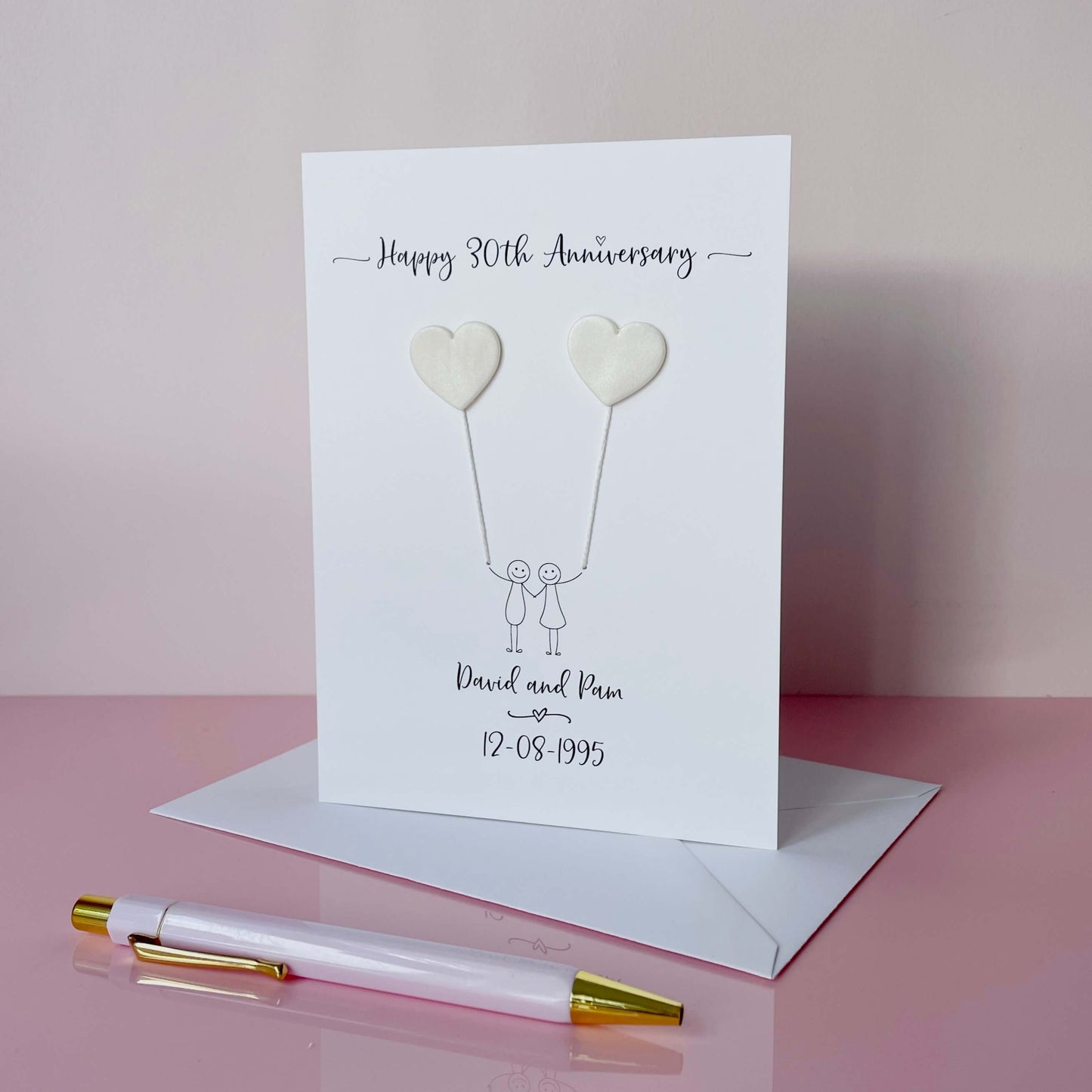 A portrait pearl anniversary card features two pearlised polymer clay hearts attached by cotton yarn, creating the illusion of balloons being held by a hand-drawn couple. The top of the card reads “Happy 30th Anniversary,” while the names and wedding date are printed at the bottom. 