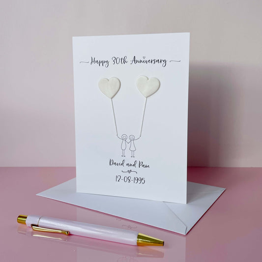 A portrait pearl anniversary card features two pearlised polymer clay hearts attached by cotton yarn, creating the illusion of balloons being held by a hand-drawn couple. The top of the card reads “Happy 30th Anniversary,” while the names and wedding date are printed at the bottom. 