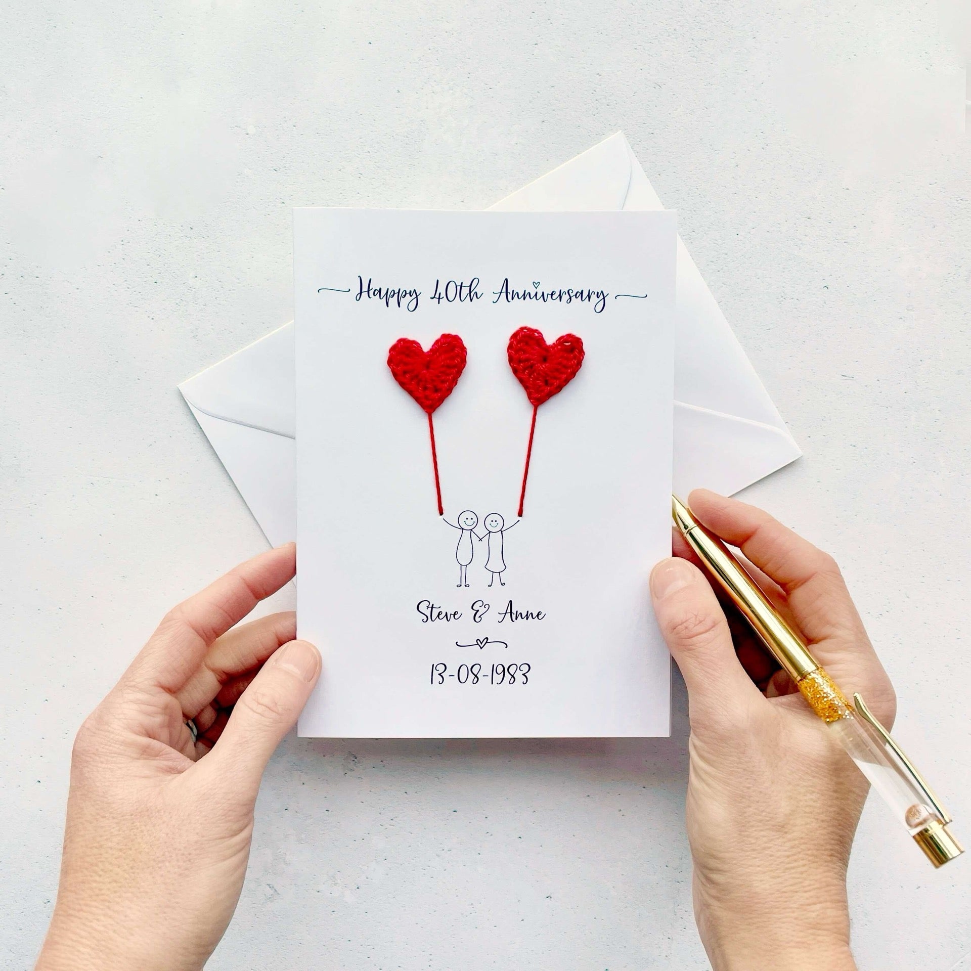 A white card featuring a line drawn couple holding hands and each holding a crochet heart shaped balloon which is made using red coloured cotton yarn. Happy 40th Anniversary is printed at the top of the card and the couples names and wedding date are printed at the bottom. 