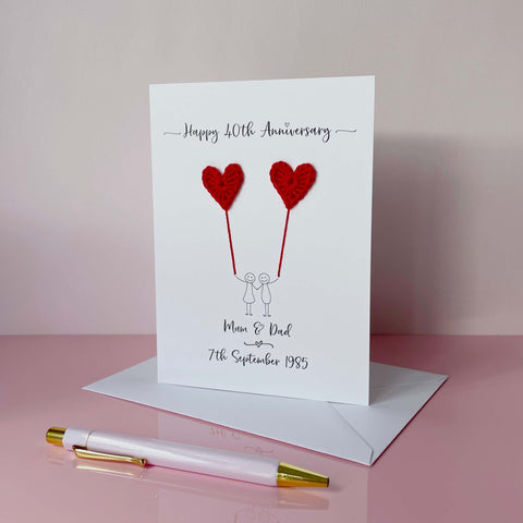 A white card featuring a line drawn couple holding hands and each holding a crochet heart shaped balloon which is made using red coloured cotton yarn. Happy 40th Anniversary is printed at the top of the card and the couples names and wedding date are printed at the bottom. 