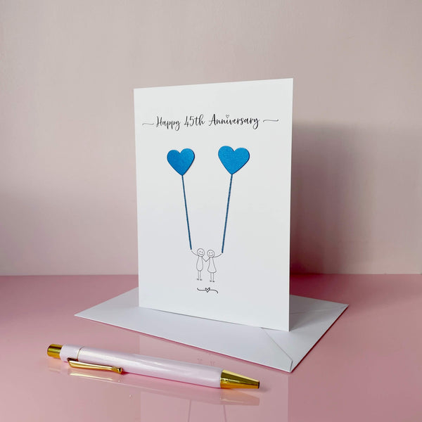 Anniversary card with blue heart balloons and two people, on a pink surface with a pen.