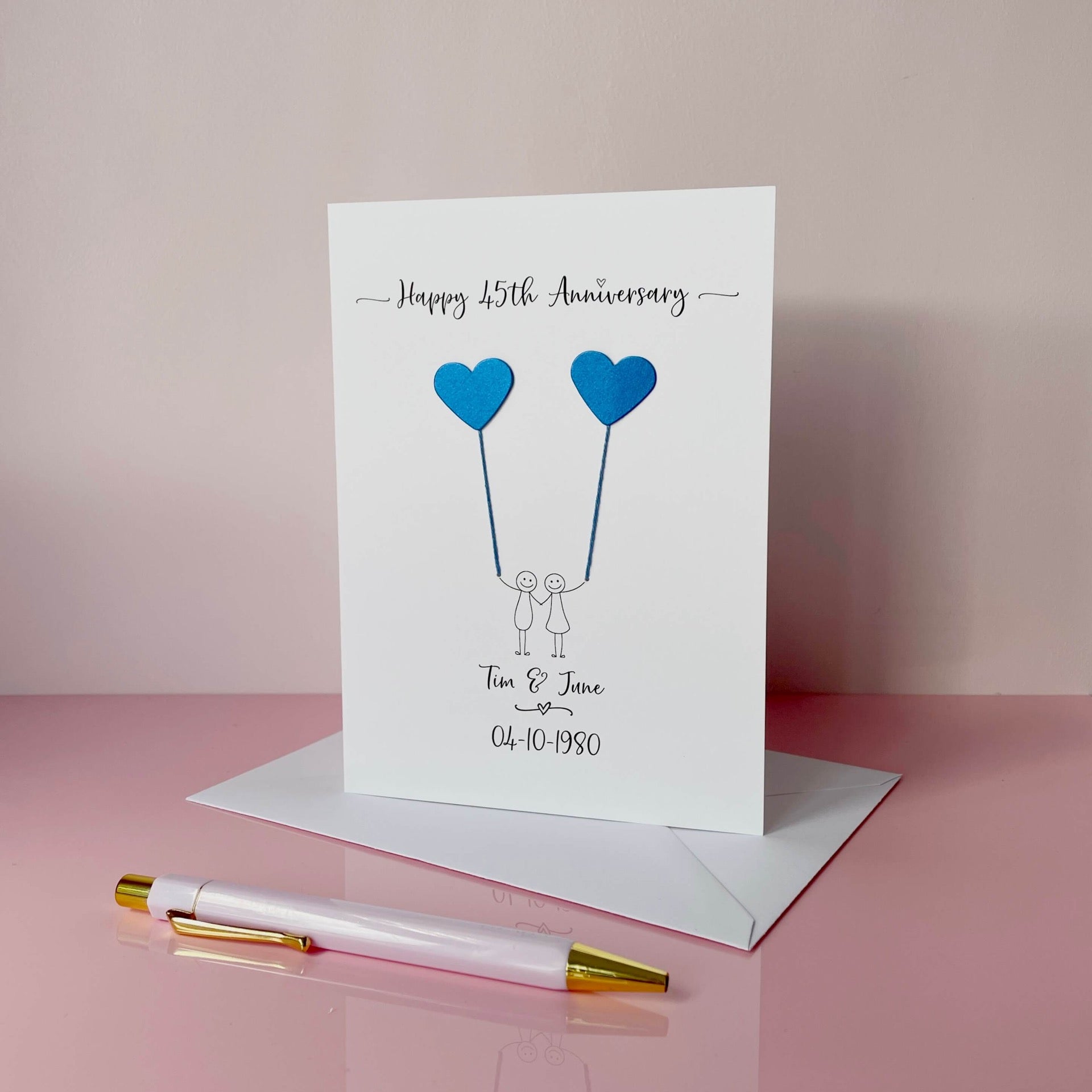 A white card featuring a line drawn couple holding hands and each holding a heart shaped balloon which is made using blue pearlised card. Happy 45th Anniversary is printed at the top of the card and the couples names and wedding date are printed at the bottom. 