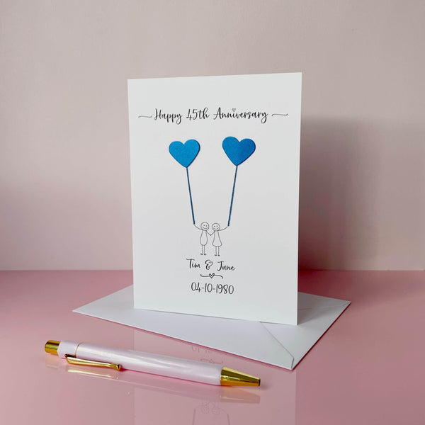 A white card featuring a line drawn couple holding hands and each holding a heart shaped balloon which is made using blue pearlised card. Happy 45th Anniversary is printed at the top of the card and the couples names and wedding date are printed at the bottom. 