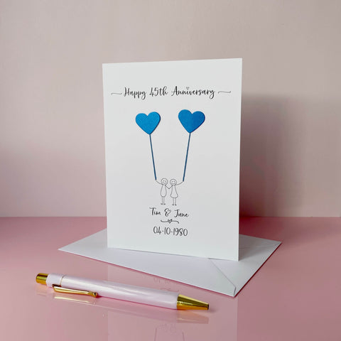 A white card featuring a line drawn couple holding hands and each holding a heart shaped balloon which is made using blue pearlised card. Happy 45th Anniversary is printed at the top of the card and the couples names and wedding date are printed at the bottom. 