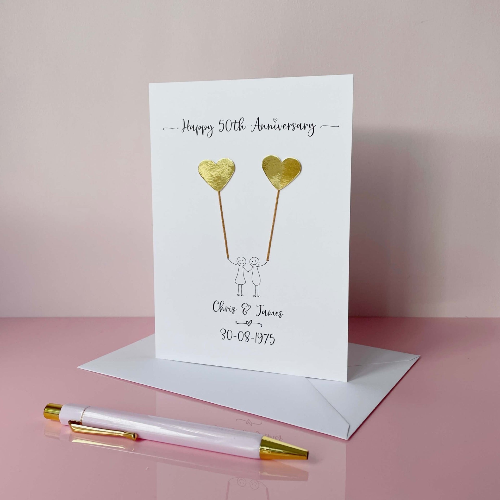 The image shows a beautiful handmade anniversary card with mirror gold card hearts, these are attached by cotton yarn, resembling balloons being held by a hand-drawn couple underneath. The text on the card reads "Happy 50th Anniversary" at the top, followed by the couples names and wedding date at the bottom. 