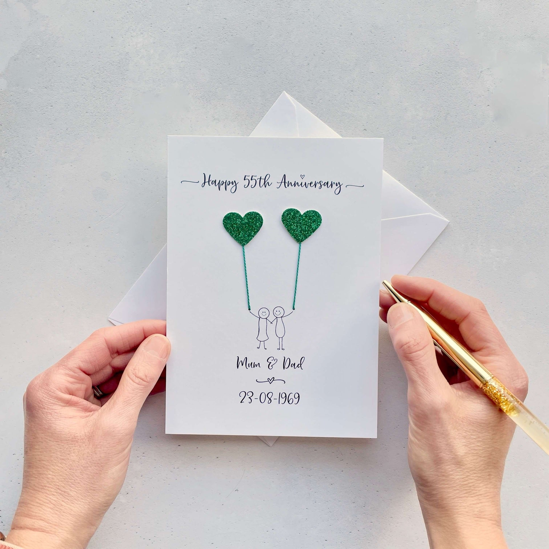 A portrait card with glitter green hearts attached by cotton yarn, resembling balloons being held by a hand-drawn couple underneath. Happy 55th Anniversary is printed at the top, followed by their names and wedding date at the bottom of the card.