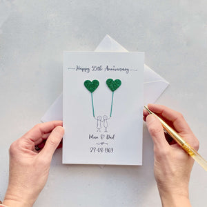 A portrait card with glitter green hearts attached by cotton yarn, resembling balloons being held by a hand-drawn couple underneath. Happy 55th Anniversary is printed at the top, followed by their names and wedding date at the bottom of the card.