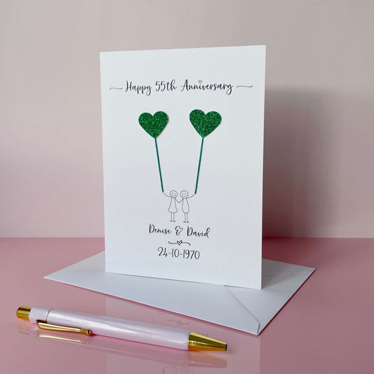 A portrait card with glitter green hearts attached by cotton yarn, resembling balloons being held by a hand-drawn couple underneath. Happy 55th Anniversary is printed at the top, followed by their names and wedding date at the bottom of the card.