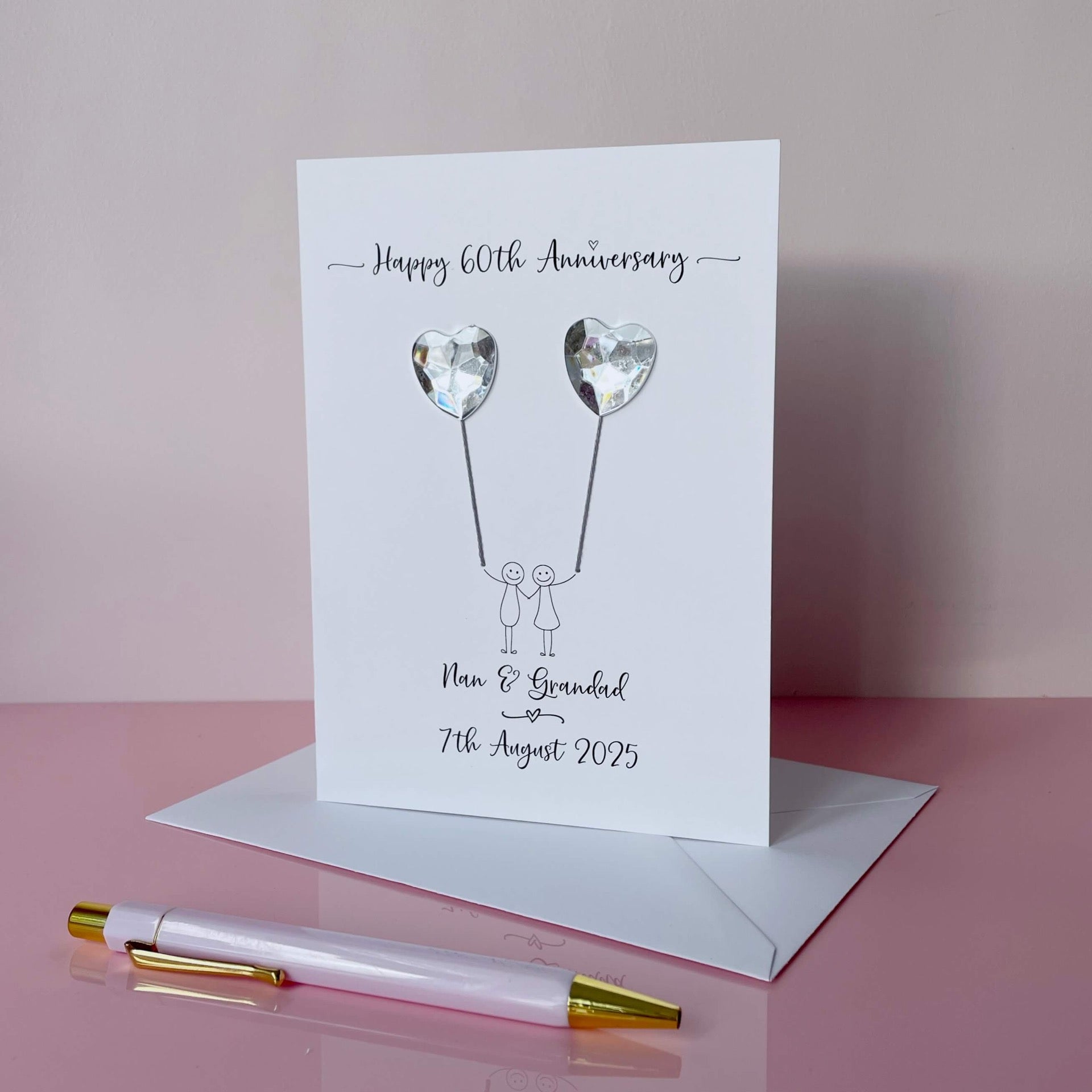 Handmade 60th anniversary card embellished with 2 heart shaped plastic gems, both attached by cotton yarn, resembling balloons being held by a couple underneath. Happy 60th Anniversary is printed at the top, the couples names/ wedding date at the bottom.
