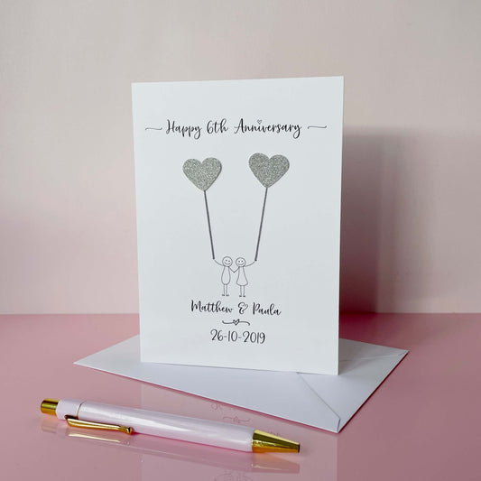 A white card with a simple line drawn couple on the front. They are each holding a sparkly grey card heart. Happy 6th anniversary is printed at the top of the card and the couples names and wedding date is printed at the bottom.