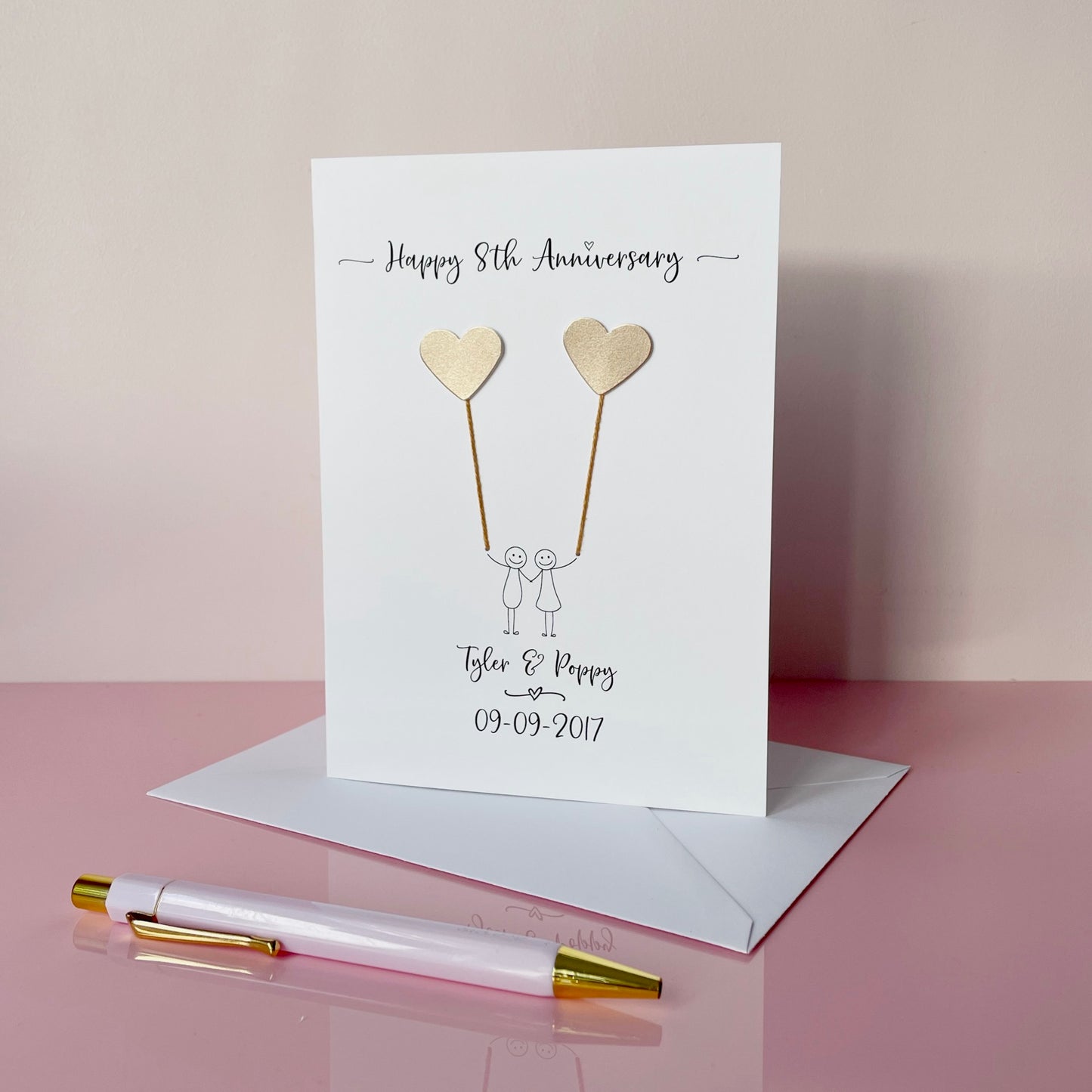 8th Anniversary card with hearts and couple illustration on a pink surface with a pen.