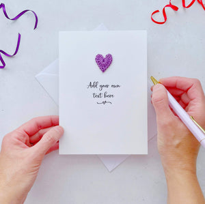 A white card with 'Add your own text here' printed in black cursive text. A purple crochet heart has been added above the text. 