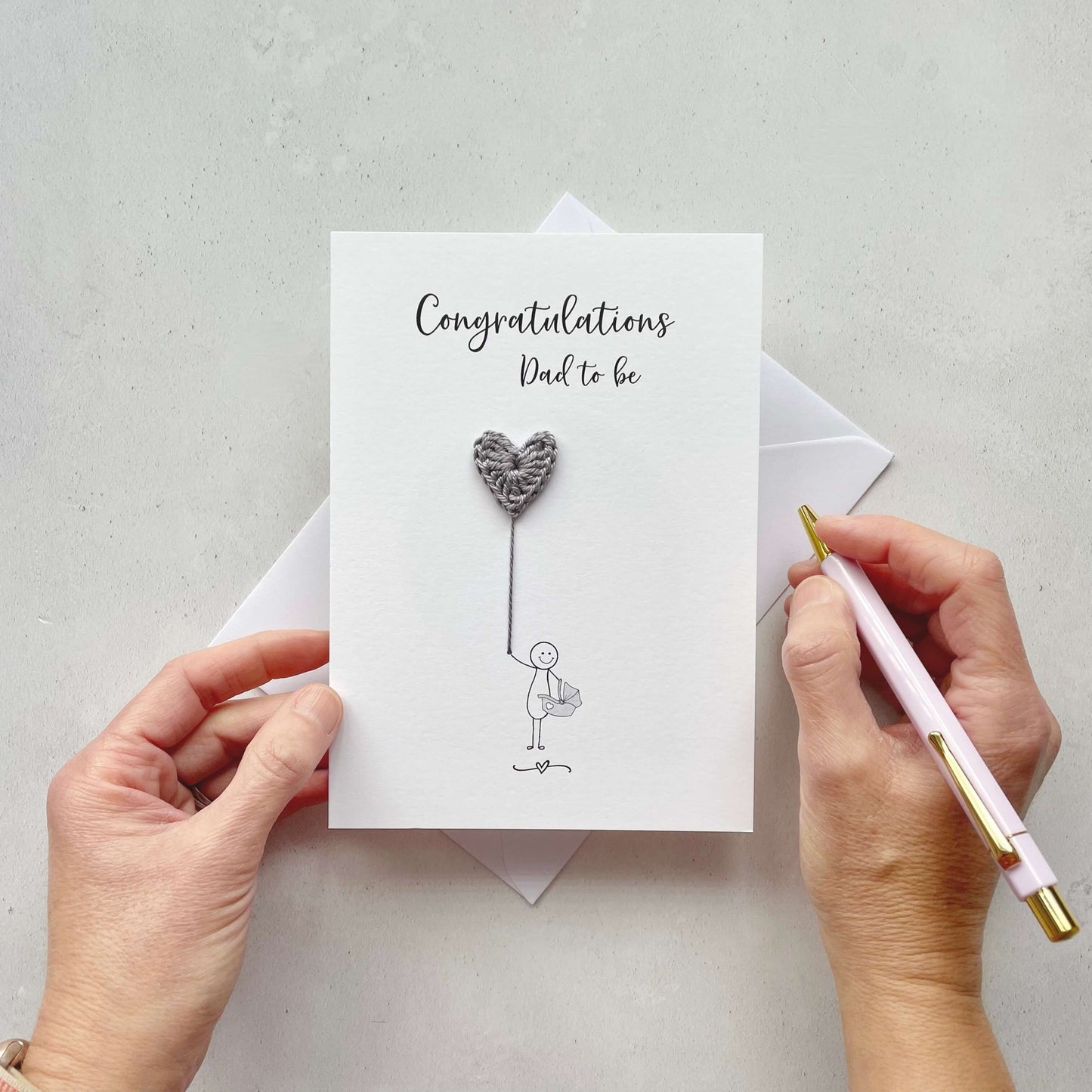 A card featuring a line drawn male, holding a pale blue crochet heart balloon. ‘To a special Daddy to be’ is printed at the top of the card in black cursive text .The character is drawn holding a baby car carrier and the word ‘Congratulations!’ is printed below him.