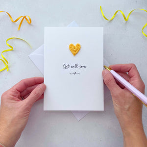 A white portrait-style card featuring the word ‘Get well soon’ in elegant black cursive at the center. Below the text, a small black squiggle and heart design adds a playful touch. Above the word, a handmade yellow crochet heart is carefully attached, providing a pop of colour and texture.