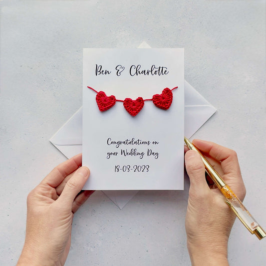 A handmade personalised wedding card with a string of 3 red crochet hearts across the front. Above the hearts are the two names and at the bottom of the card is 'Congratulations on your wedding day' and the wedding date. 