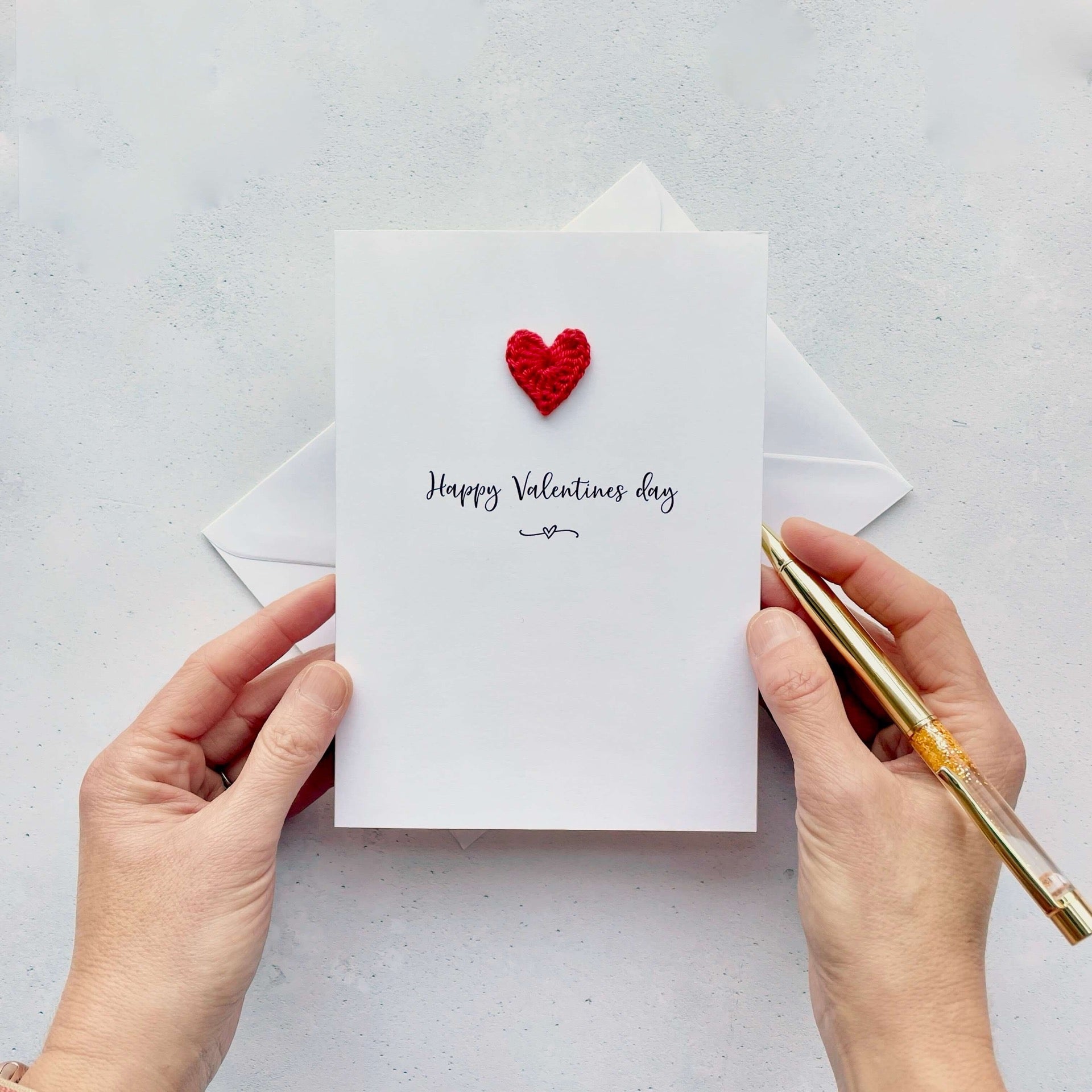 A white card with a mini red crochet heart  on it. Under the heart is printed "Happy Valentine's Day' in black cursive text.