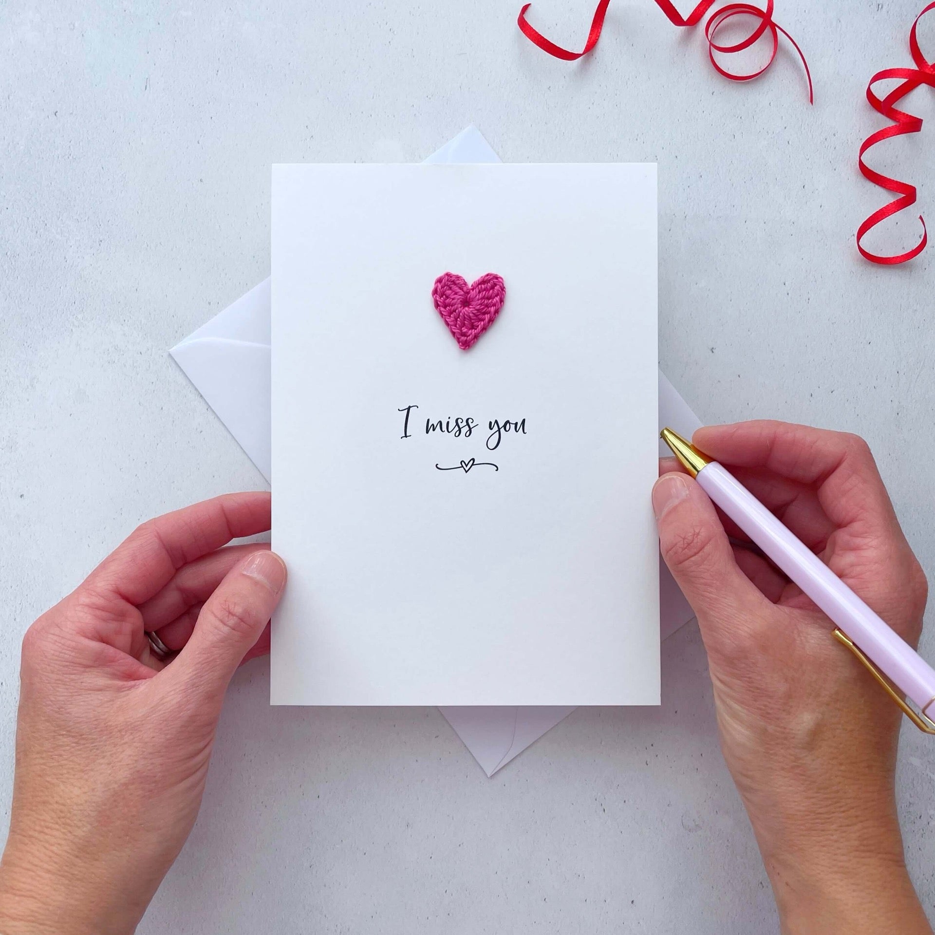 A white portrait-style card featuring the words ‘I miss you’ in elegant black cursive at the center. Below the text, a small black squiggle and heart design adds a playful touch. Above the words, a handmade bright pink crochet heart is carefully attached, providing a pop of colour and texture.