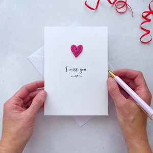 A white portrait-style card featuring the words ‘I miss you’ in elegant black cursive at the center. Below the text, a small black squiggle and heart design adds a playful touch. Above the words, a handmade bright pink crochet heart is carefully attached, providing a pop of colour and texture.