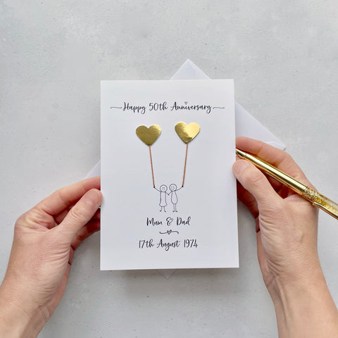 The image shows a beautiful handmade anniversary card with mirror gold card hearts, these are attached by cotton yarn, resembling balloons being held by a hand-drawn couple underneath. The text on the card reads "Happy 50th Anniversary" at the top, followed by the couples names and wedding date at the bottom. The card is set on a light background, and the person's hands are seen holding a pen and gently holding the card, giving the image a sense of care and personalisation.